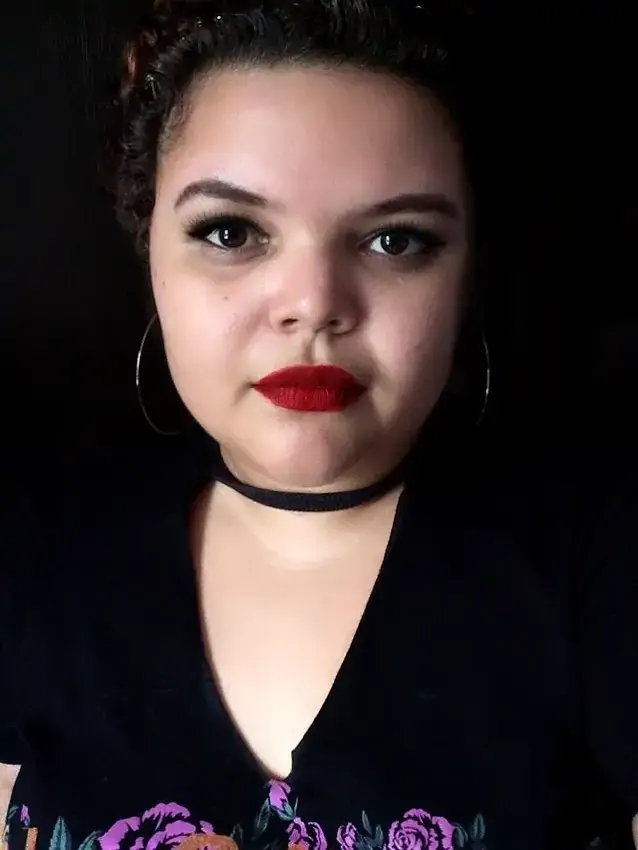 Foto de Niedja Araujo, uma mulher jovem com cabelos cacheados e batom vermelho